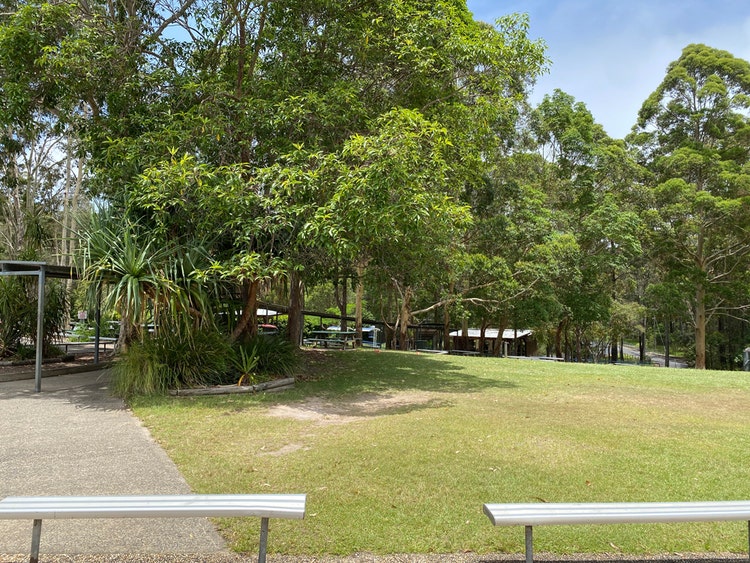 Two aluminium bench seats with tall trees and a green playground in the background.