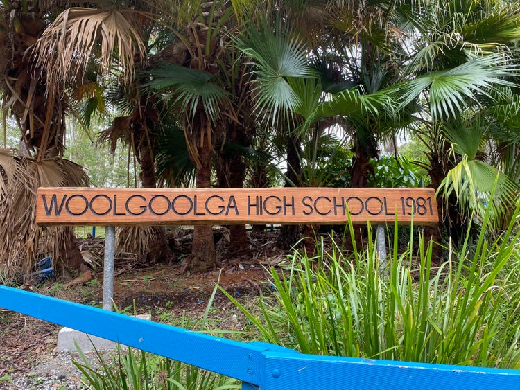 Woolgoolga High School wooden sign underneath large cabbage palm leaves